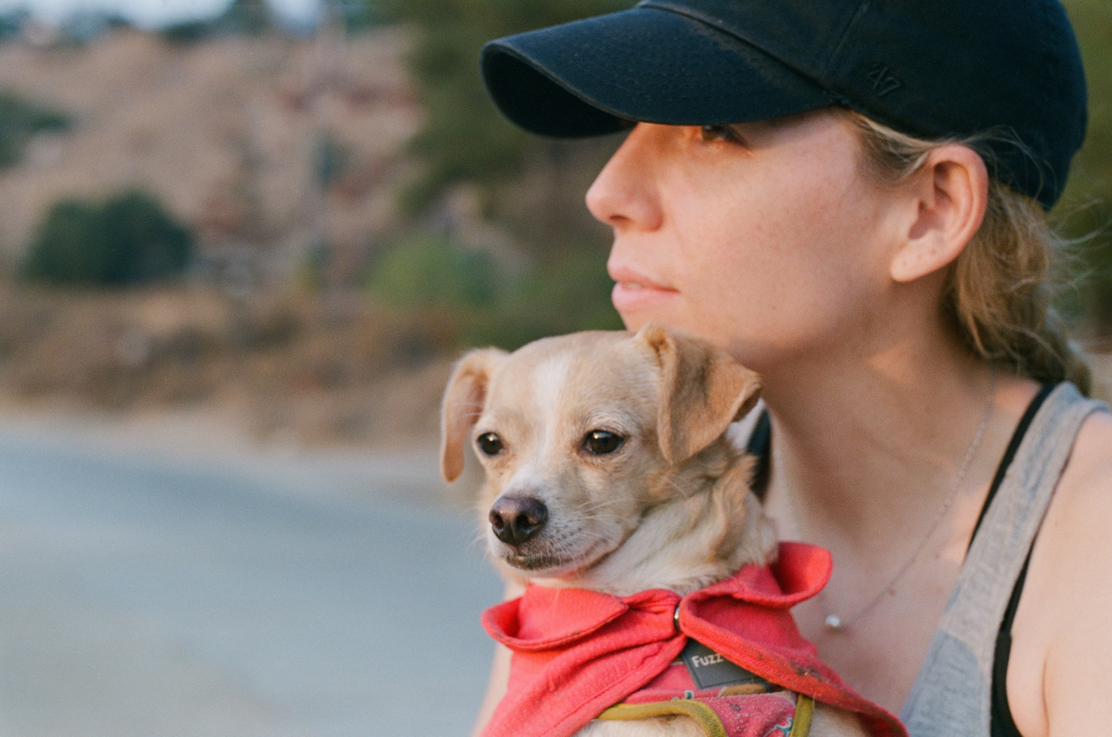 Woman wearing a black cap holds a small tan dog in a red bandana outdoors.