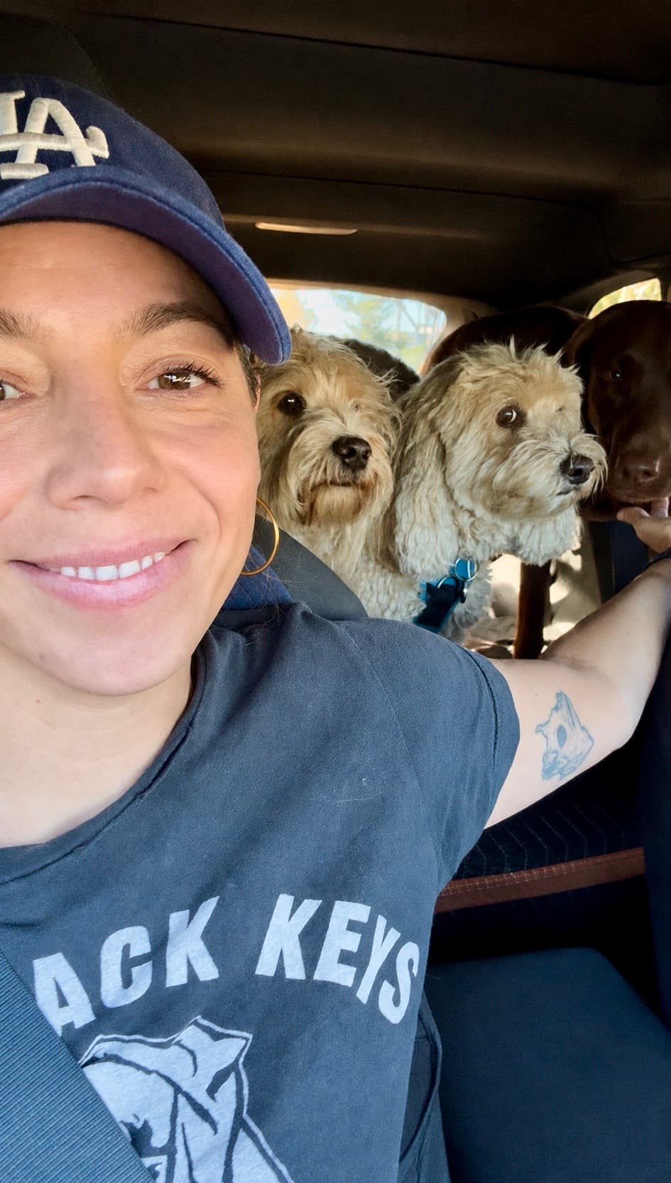 Smiling woman in a Dodgers cap takes a selfie with three dogs in a car.